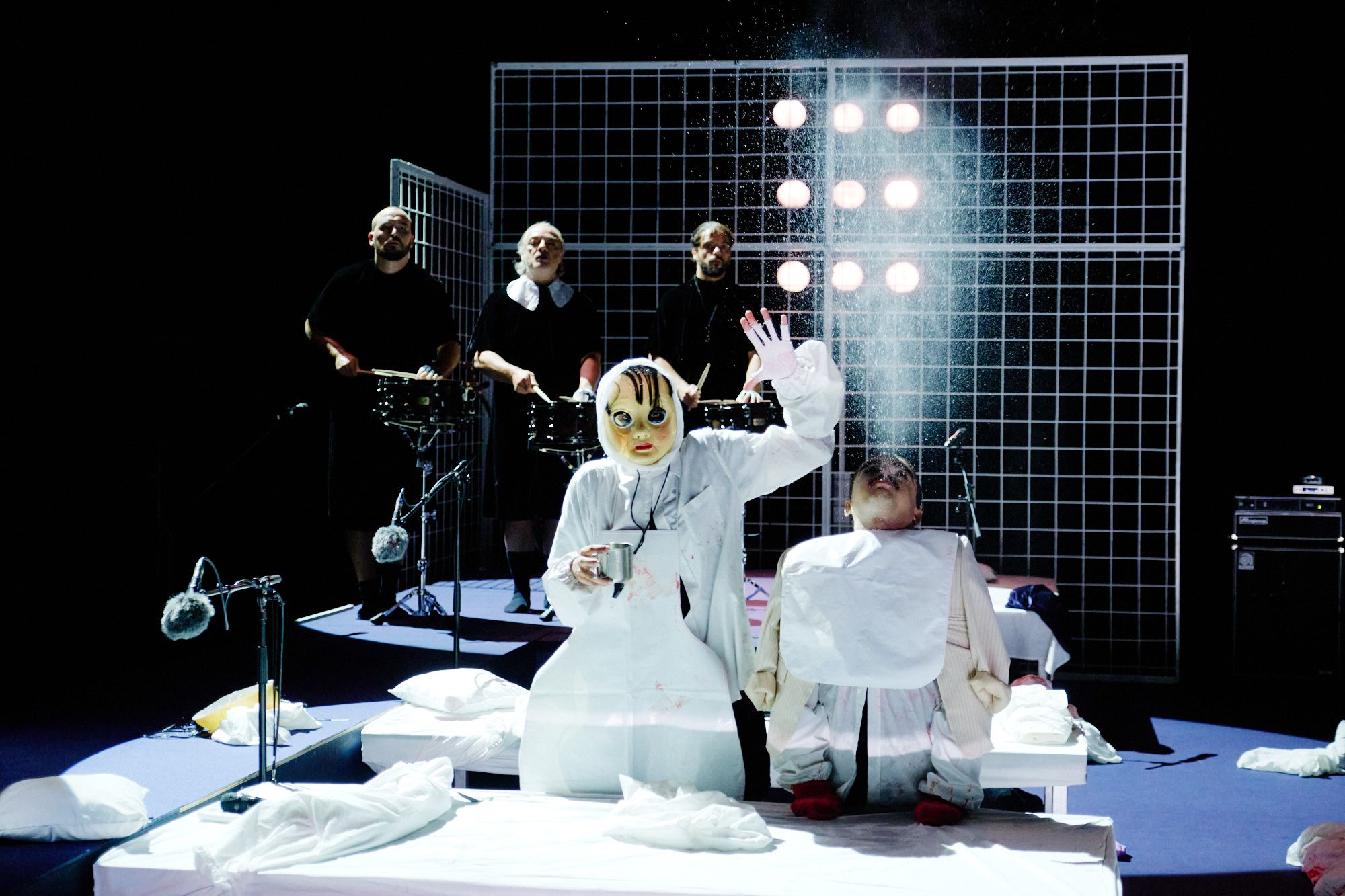 Eine Szene aus NÔT: Zwei Performer*innen, die zwischen zwei Betten auf einer dunklen Bühne sitzen. Der*die Performer*in links ist maskiert und in weiß gekleidet. In der einen Hand ist eine Tasse zu sehen, die linke Hand in der Luft. Daneben eine kleine Person, die mit nach hinten geworfenem Kopf Wasser in die Luft pustet. Im Hintergrund drei schwarz gekleidete Musiker*innen, die vor einer Gitterwand stehen, an der Scheinwerfer zu sehen sind.