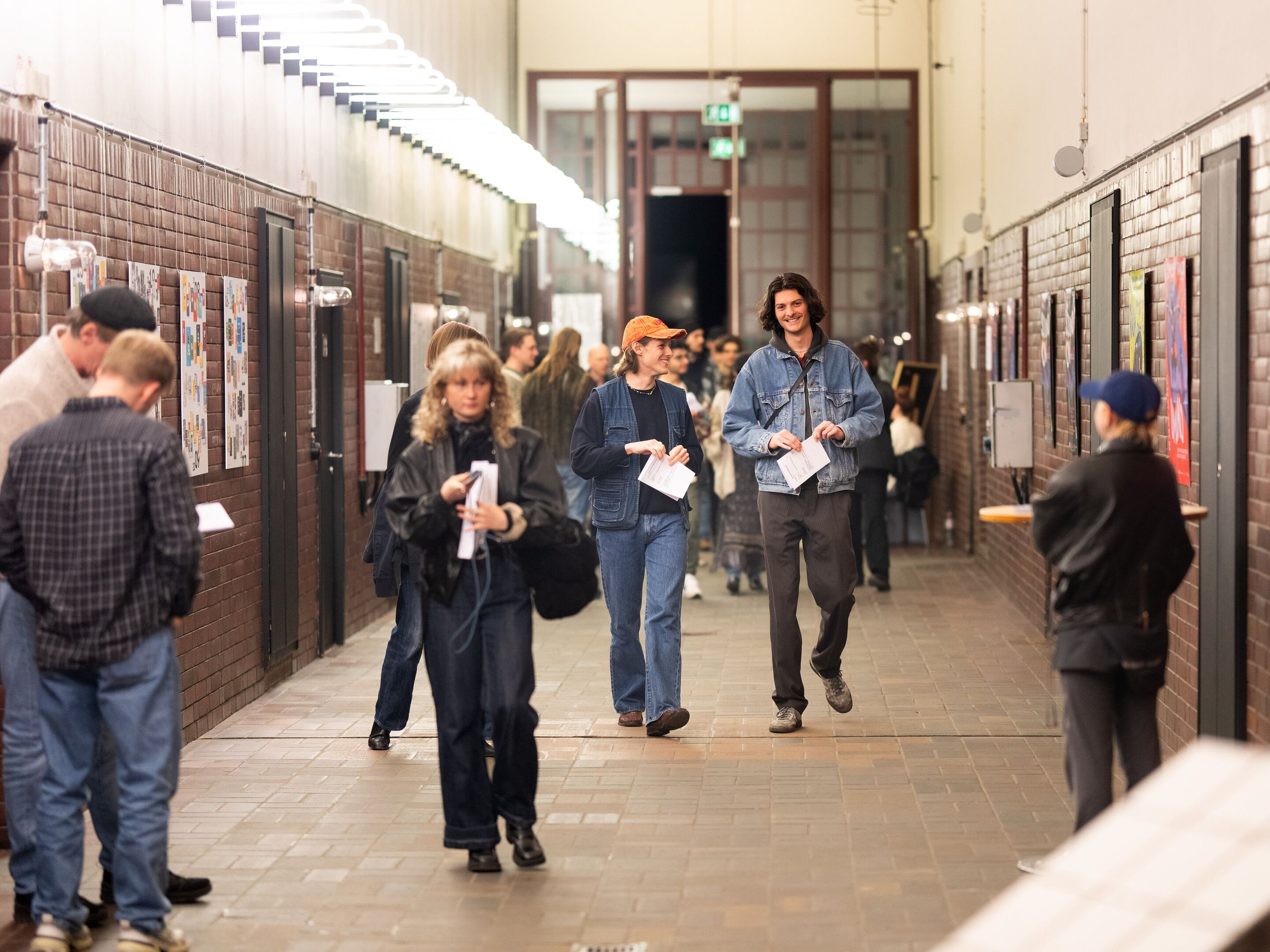 A group of young people are walking along a long corridor. They are laughing and engrossed in conversation. Many people are standing around them.