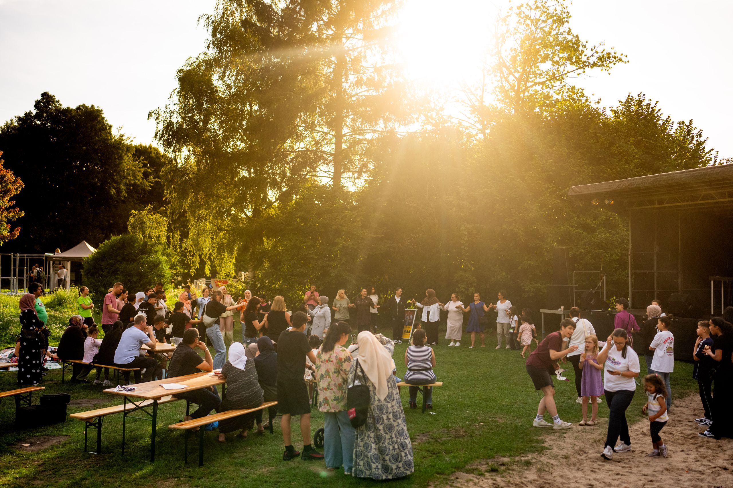 Tables and beer benches are set up on a meadow. Numerous people gather. They stand in a large circle, hold hands and dance together.