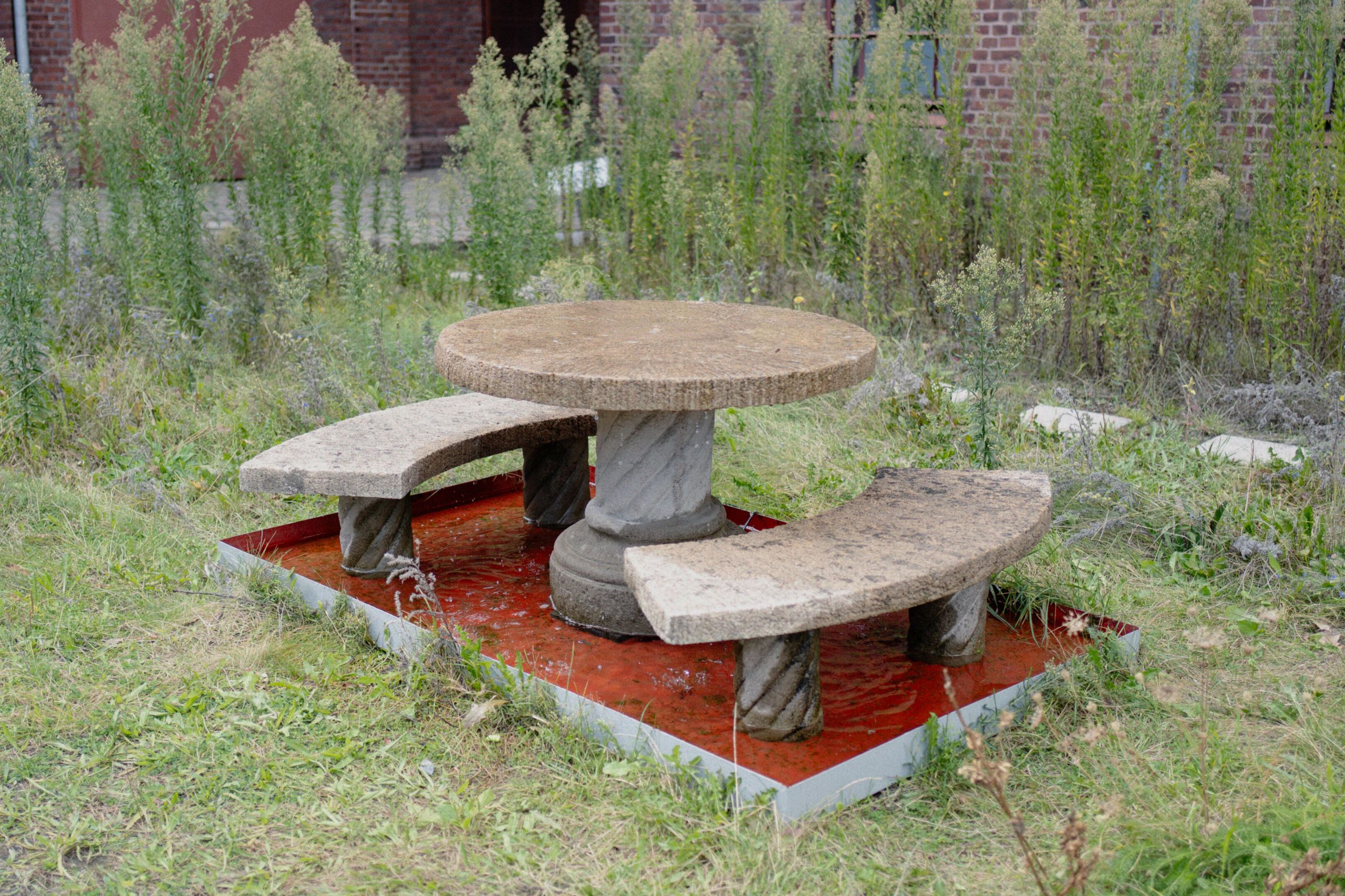 Eine Brunneninstallation auf der begrünten Fläche vor PACT Zollverein. Es handelt sich um einen runden Tisch aus Stein, aus dessen Mitte Wasser sprudelt. Drumherum zwei Bänke. Tisch und Bänke stehen in einem roten Becken.