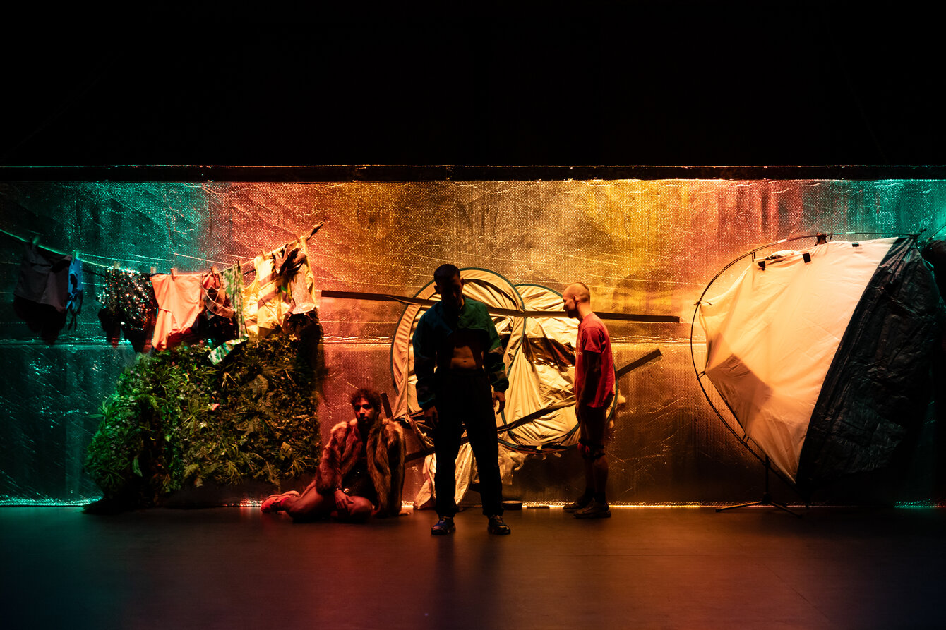 Drei Personen stehen in buntem Bühnenlicht vor einer Wand aus reflektierender Folie und diversen Requisiten