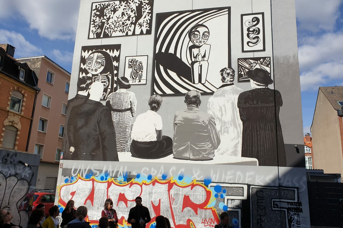 A group of people is gathered in front of a house facade, on which there is a large picture in street art style.