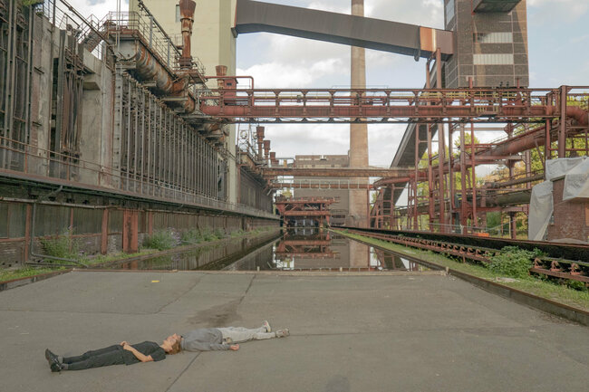 Zwei Perosnen liegen auf dem Betonboden vor der Kokerei der Zeche Zollverein