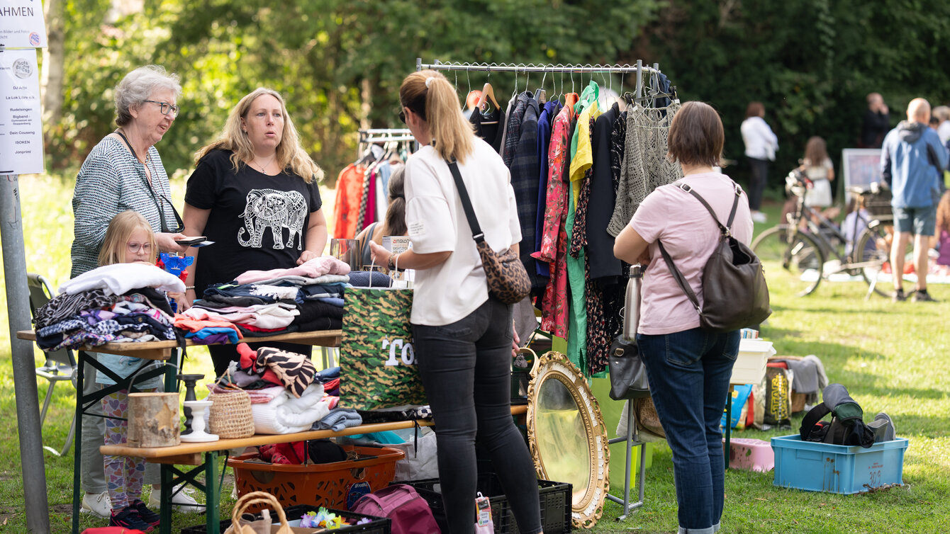 Eine Gruppe von Personen verschiedenen Alters steht um einen Flohmarktstand verteilt. Auf Tischen, Bänken und Ständern sind zahlreiche Kleidungsstücke und Wohnutensilien ausgebreitet.
