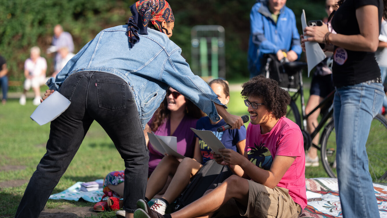 Eine Gruppe junger Menschen sitzt auf einer Wiese. Sie halten einen Liedtext vor sich und singen gemeinsam. Eine weitere Person wendet sich der Gruppe zu und hält für sie ein Mikrofon.