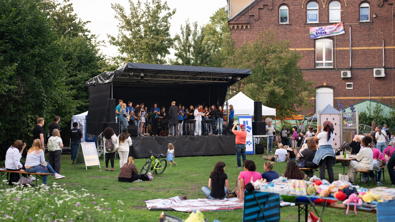 Eine Gruppe von Menschen versammelt sich auf einer Wiese. Sie stehen um eine Bühen verteilt, auf welcher ein Schulorchester spielt.