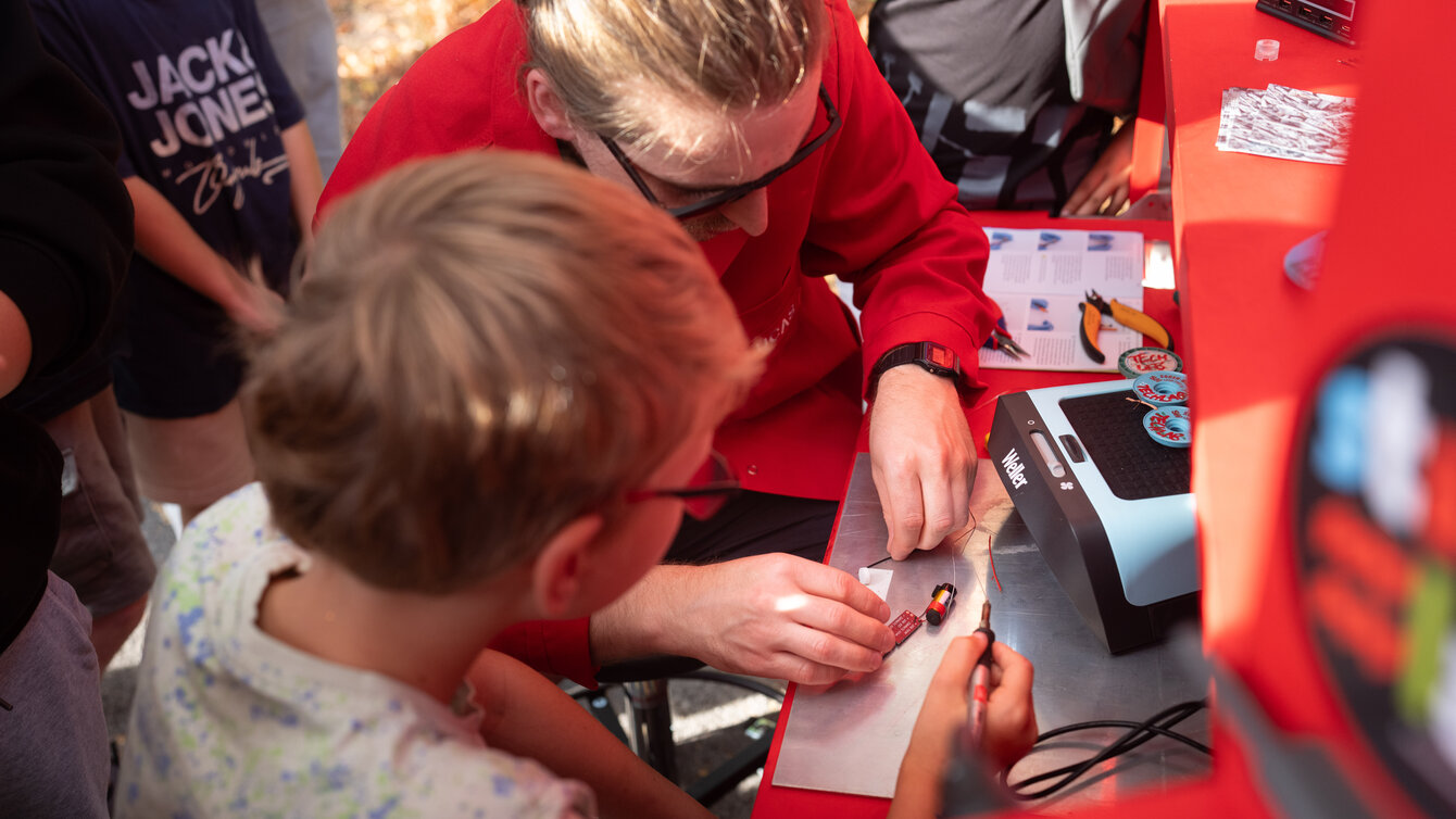 Eine Gruppe von Kindern steht um eine Werkbank verteilt. Gemeinsam mit einer Person in rotem Overall löten sie Elektroteile.