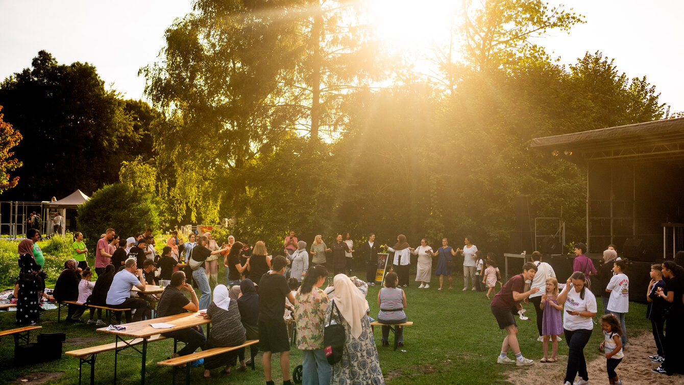 Auf einer Wiese sind Tische und Bierbänke aufgestellt. Zahlreiche Menschen versammeln sich. Sie stehen in einem großen Kreis, halten sich an den Händen und tanzen gemeinsam.