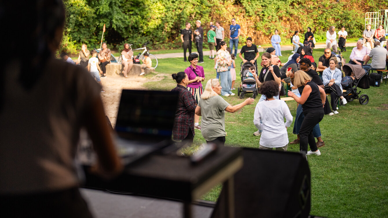 Eine Gruppe steht auf einer Wiese und tanzt gemeinsam. Um sie herum sind Menschen verteilt und beobachten das Geschehen. Im Vordergrund ist eine Person zu sehen, welche auf einer Bühne steht und Musik auflegt.