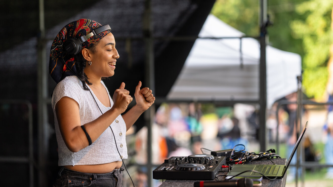 Eine Person steht auf einer Bühne. Vor ihr steht ein Pult mit einem DJ-Controller. Sie lächelt und tanzt euphorisch.