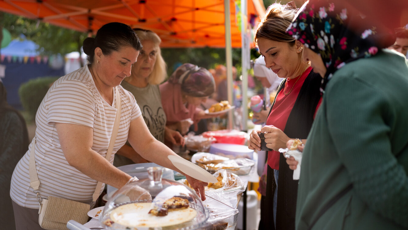 Mehrere Personen stehen an einem Essensstand. Auf einem langen gedeckten Tisch sind süße Gebäckstücke und Knabberein aufbereitet.