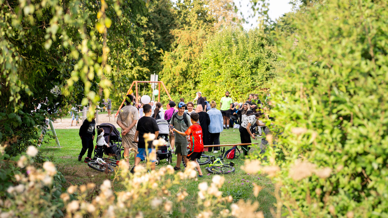 Eine Gruppe von Menschen versammelt sich auf einer Wiese. Etwas abseits der Veranstaltung steht eine Gruppe Kinder. Sie tragen Fußballtrikots und essen Süßigkeiten.