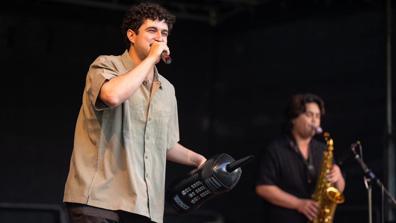 Der Rapper „Dein Couseng" steht auf einer Bühne und rappt für das Publikum. Im Hintergrund steht ein weiterer Musiker und spielt Saxophon.