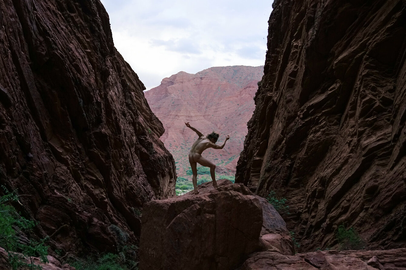 In einer Kluft steht eine Person auf einem Felsen. Sie ist nackt und nimmt eine Pose tänzerische Pose ein. Im Hintergrund ist eine Berglandschaft zu sehen, vor derer sie sich deutlich abhebt. 