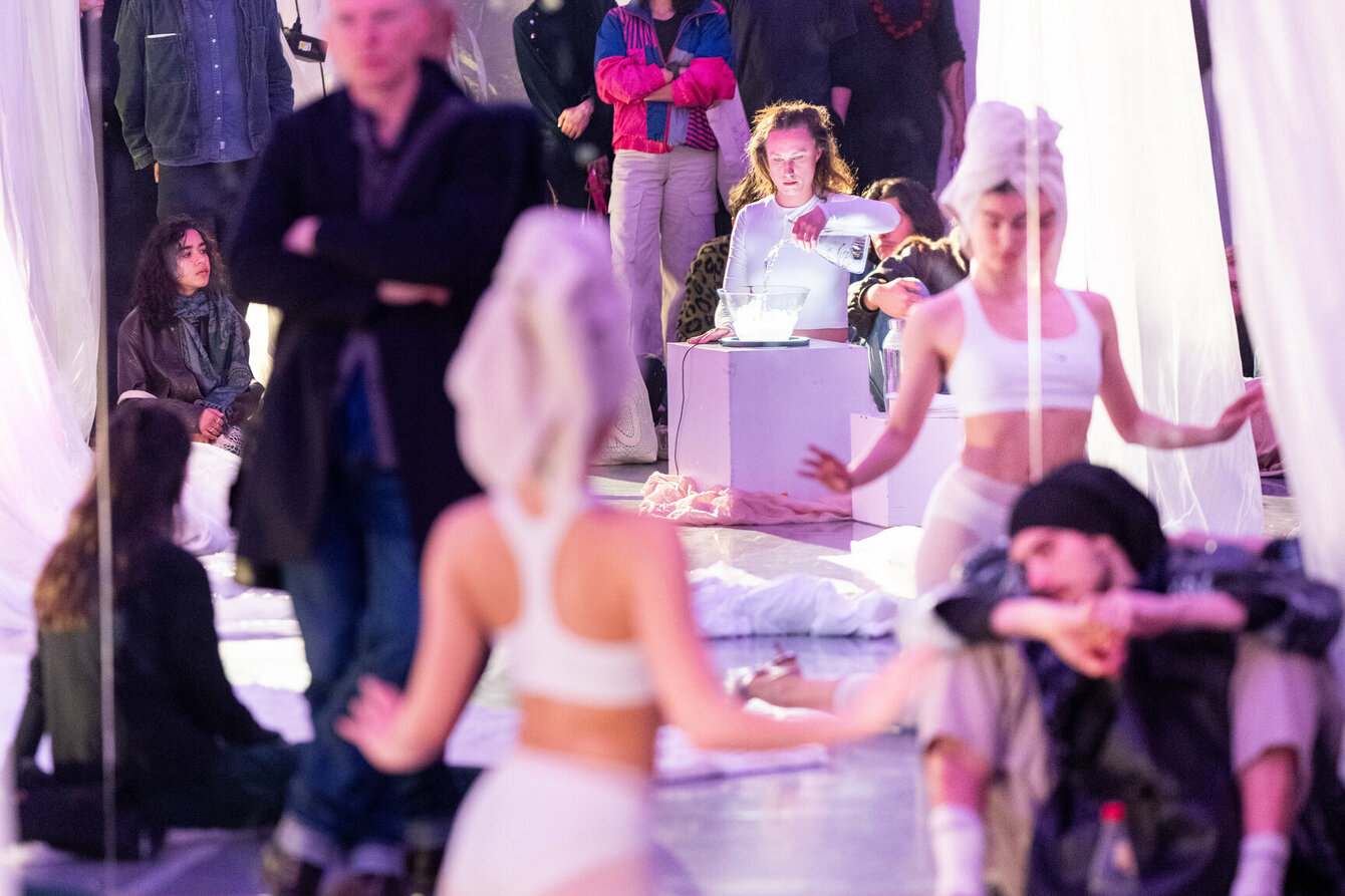 Eine Person befindet sich in einem Tanzsaal und betrachtet sich im Spiegel. Sie trägt weiße, sportliche Kleidung und Schuhe mit schmalen Absätzen. Ihre Haare sind in einem Handtuch eingewickelt. Im Spiegel ist eine weitere Person zu erkennen, welche in eine erleuchtete Schale voller Eiswürfel Wasser gießt. Die beiden Personen sind von Zuschauer:innen umgeben.