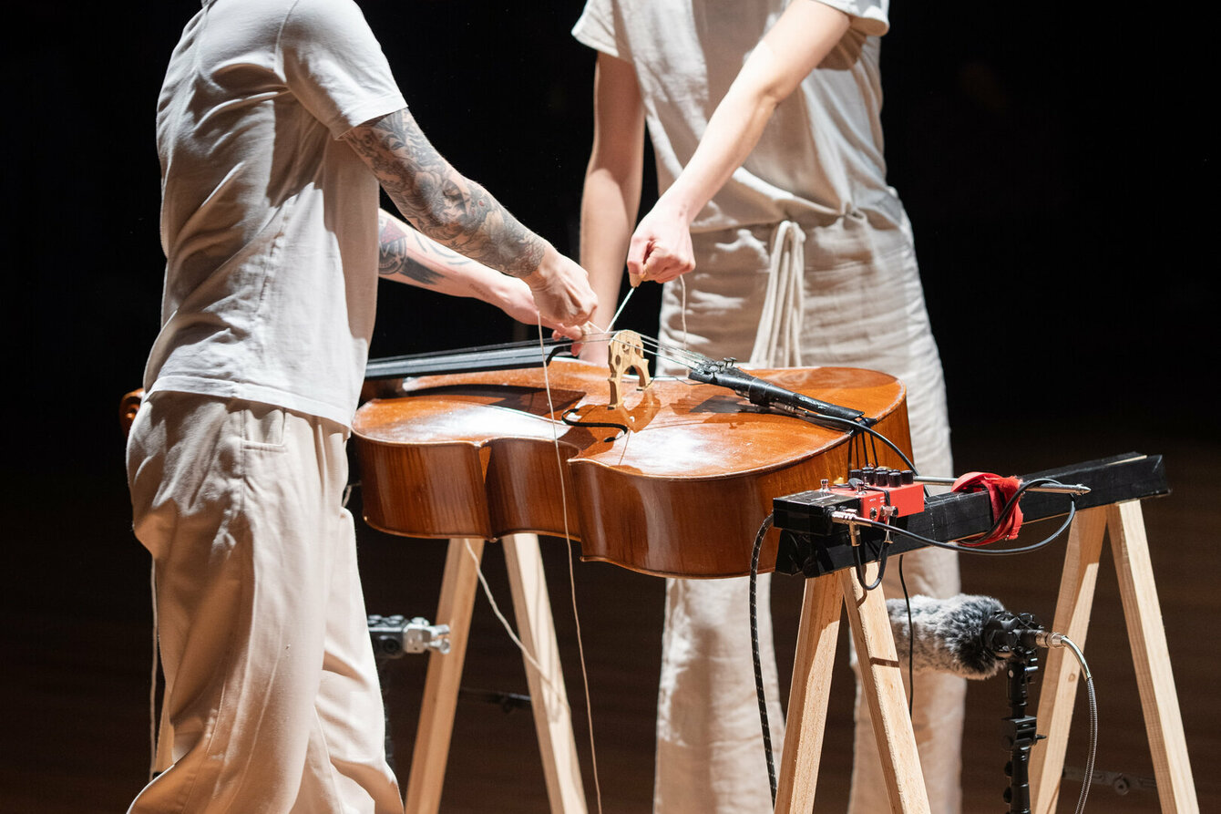 Zwei Personen stehen sich gegenüber. Sie befinden sich auf einer Bühne und werden von Scheinwerfern angestrahlt. Zwischen Ihnen befindet sich ein Cello, welches auf zwei Holzböcken befestigt, wagerecht vor ihnen liegt. Sie halten einen Faden in ihren Händen, welcher über die Saiten des Instruments streicht.