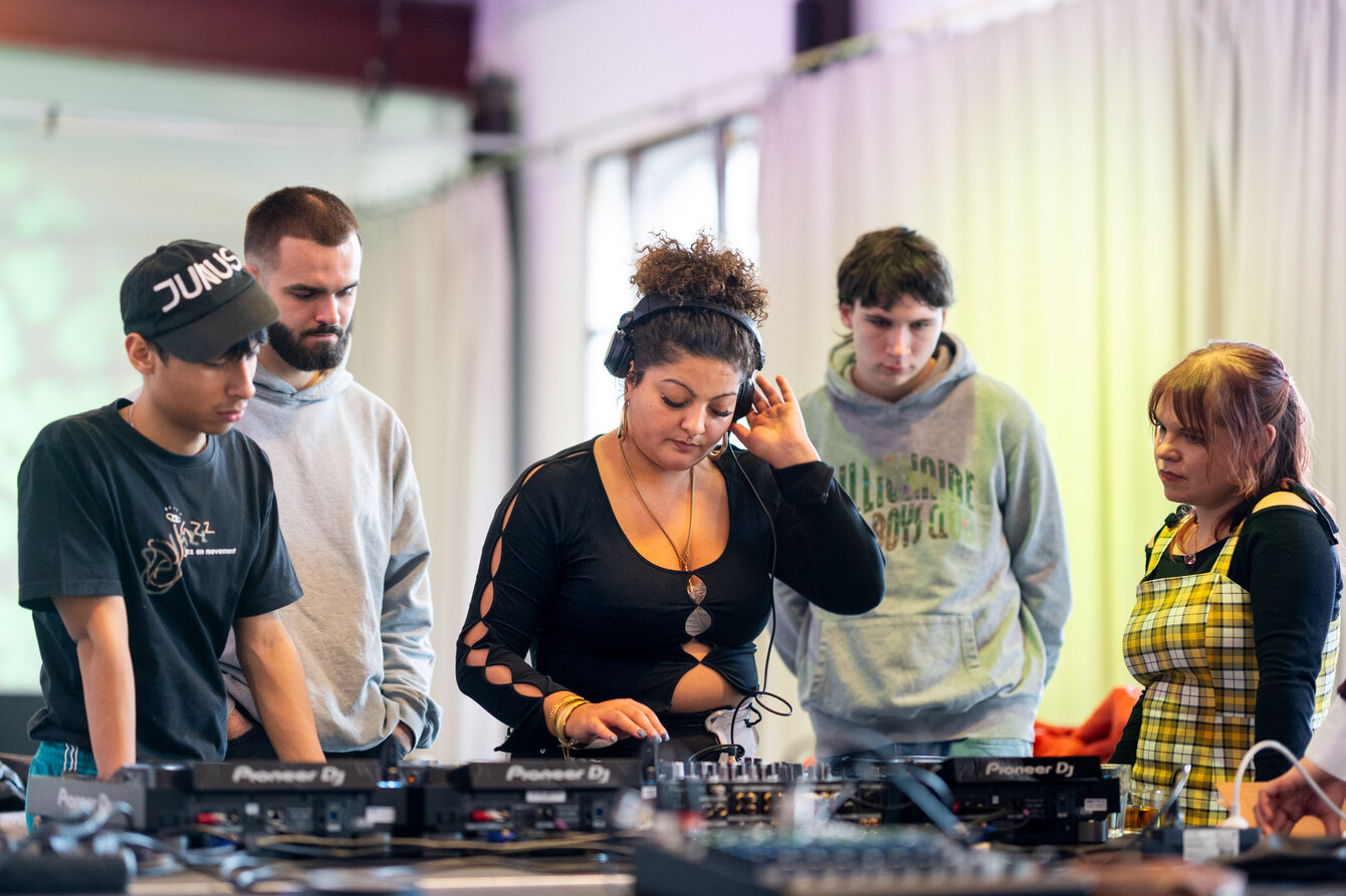 Eine Gruppe junger Menschen steht vor einem DJ Pult. Eine Person legt auf, die anderen beobachten genau, wie die Person arbeitet.