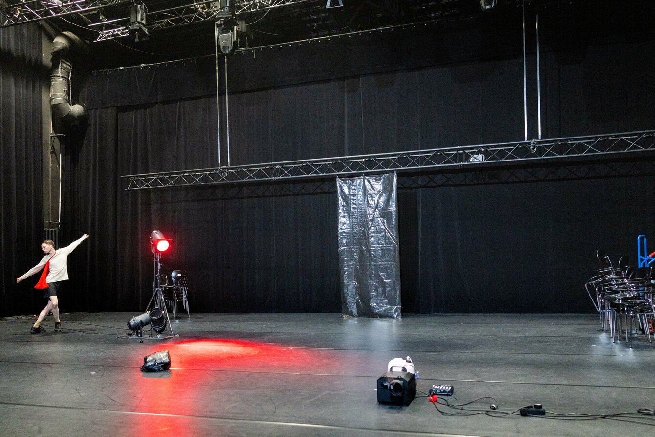 Tänzer auf einer fast leeren Bühne, auf der sich eine rote Lampe und aufeinander gestapelte Stühle befinden. 