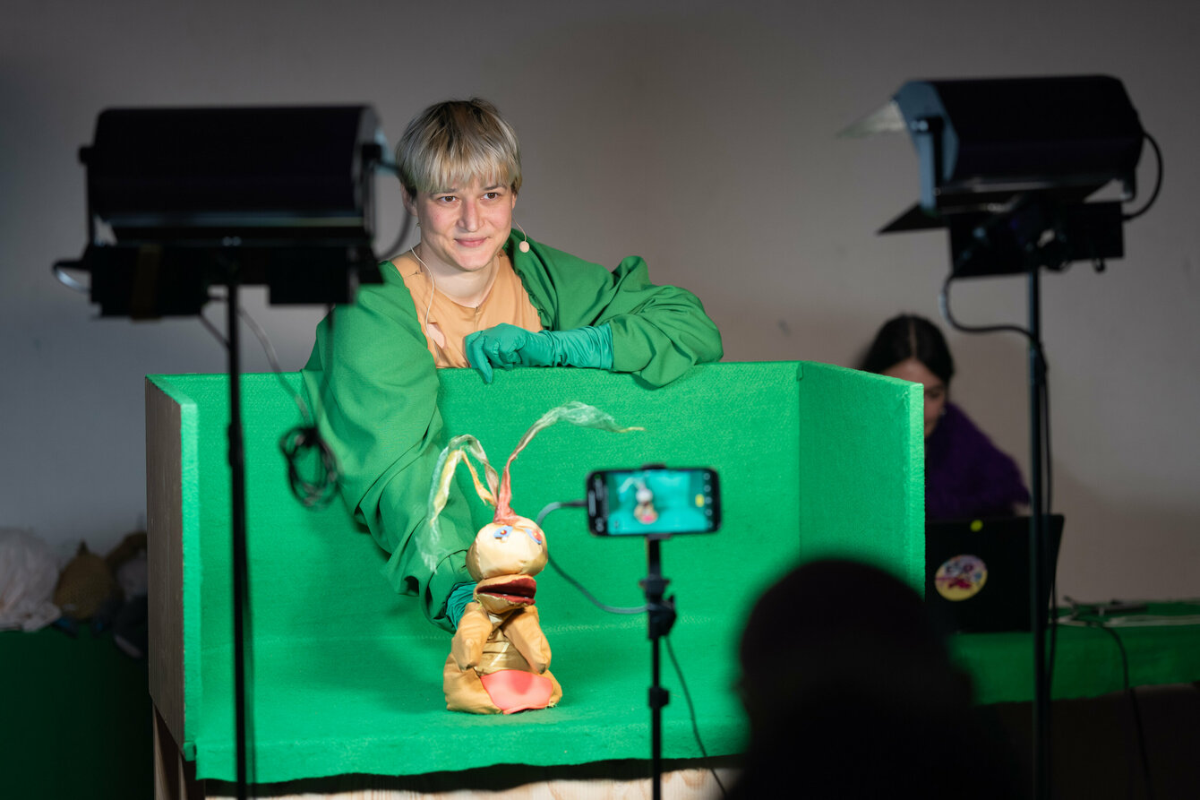 Eine Person steht vor einer Kamera. Ihre Arme und Hände sind in grünen Stoff gehüllt. Mit der rechten Hand hält sie eine Handpuppe, welche an die Form einer Zwiebelpflanze erinnert. Die Handpuppe befindet sich in einer Art Fotobox, welche in grünen Stoff gehüllt ist.