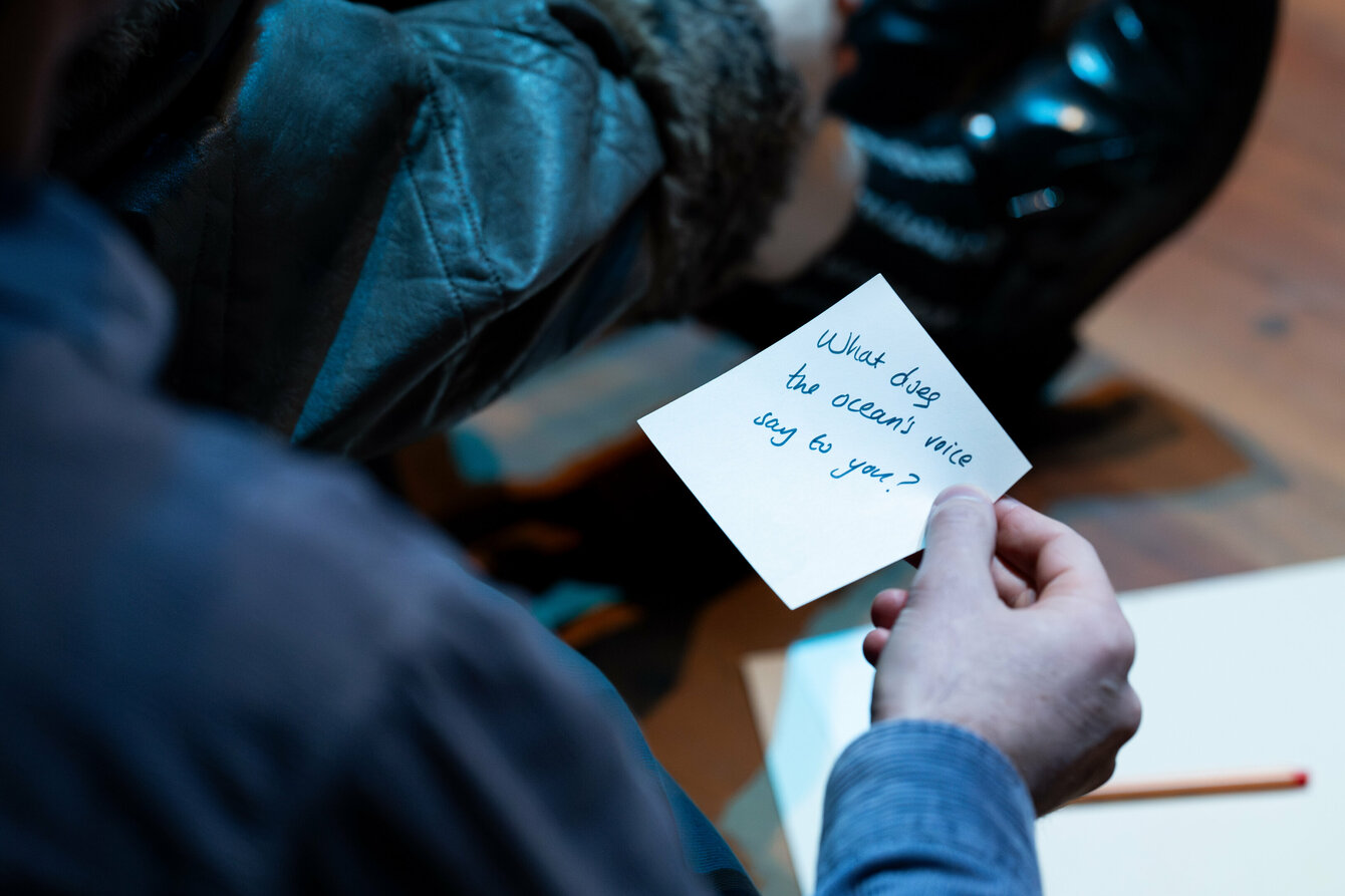 Ein:e Zuschauer:in hält einen Zettel in der Hand. In geschwungener Schrift steht auf dem Zettel: What does the Ocean's voice say to you?