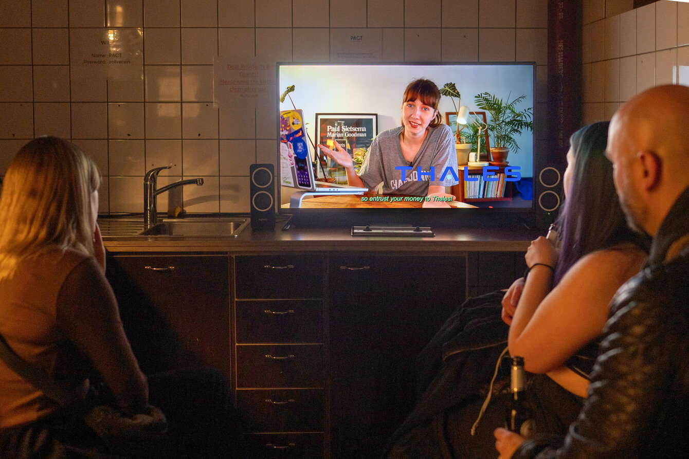 Zuschauer:innen blicken auf einen Fernseher. Auf dem Bildschirm ist eine Person zu erkennen, welche in einem Wohnzimmer sitzt und zur Kamera spricht. Auf dem Bildschirm ist das Logo des französischen Konzerns "Thales" zu erkennen.