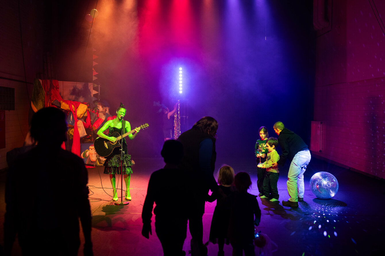 Eine Person steht auf einer Bühne. Sie trägt grüne Farbe am ganzen Körper. Sie spielt auf einer Gitarre und singt in ein Mikrofon. 