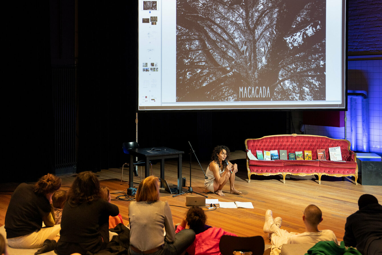Person sitzt in einem Bühnenraum auf dem Boden vor einer Gruppe von Menschen, dahinter eine Leinwand mit einem Bild eines Baumes und dem Text 'MACACADA'.