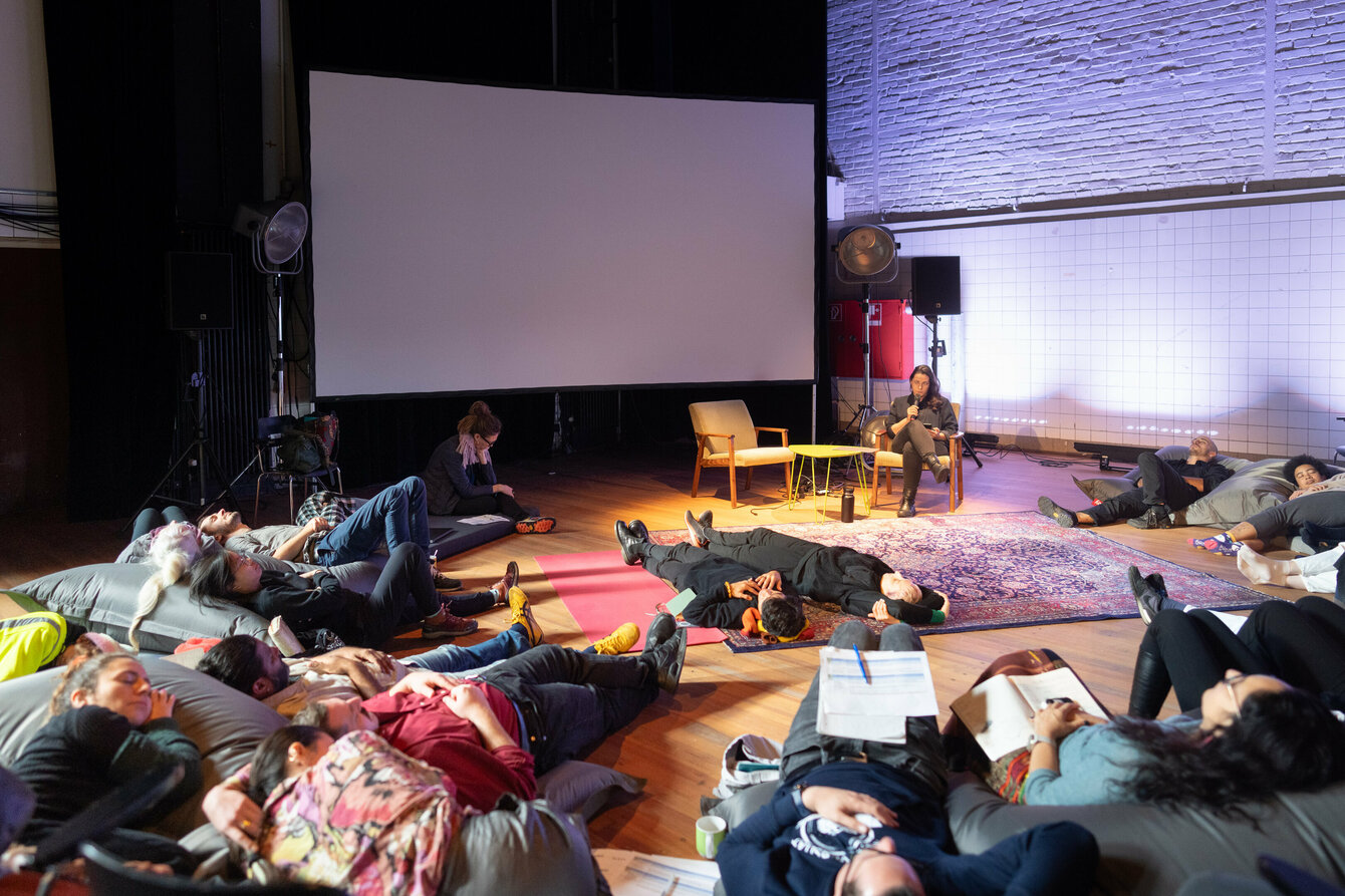 People are lying comfortably on carpets on the floor. One person sits in an armchair in front of them and gives a lecture. Next to them is a large white movie screen.