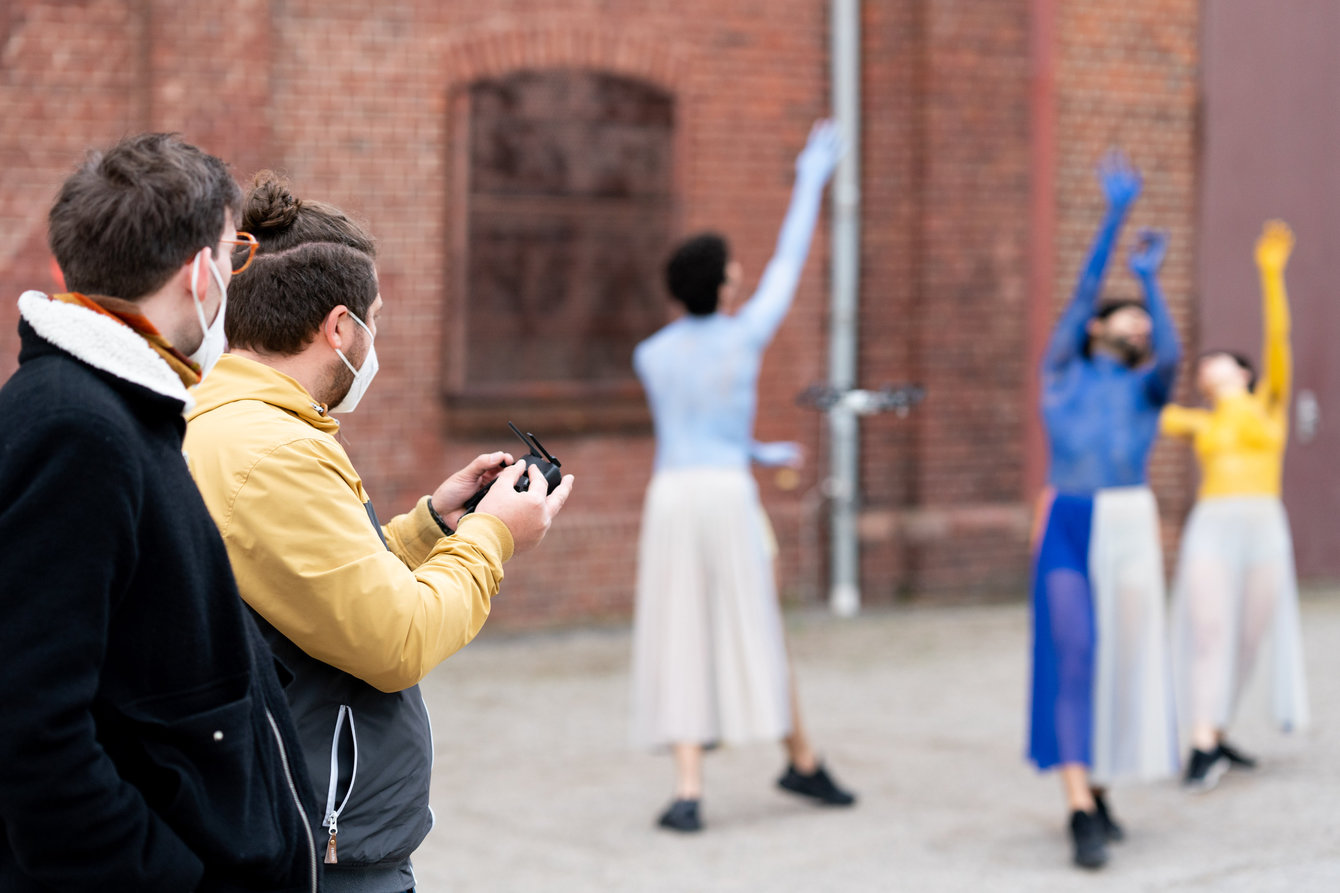 Der Choreograph schaut dem Dronen-Operator beim Filmen einer Gruppe von Tänzer:innen vor dem PACT Gebäude auf dem Zollverein-Gelände über die Schulter