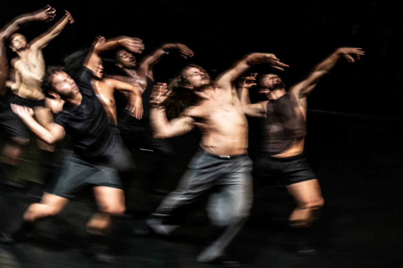 A scene from the performance F*cking Future: The image was taken with motion blur and shows a group of dancers dancing shirtless with their arms in the air on a darkened stage.