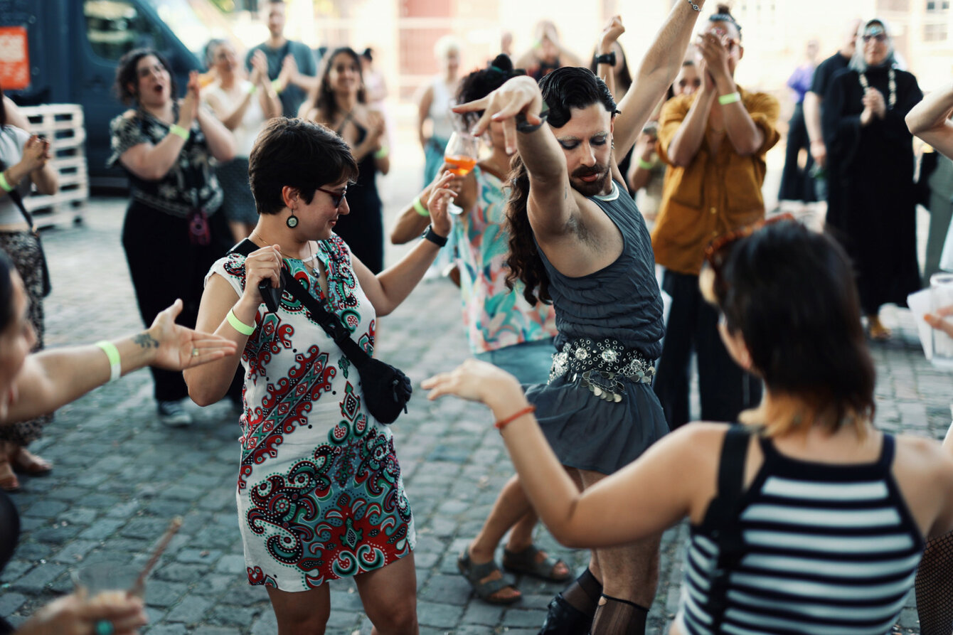 Eine Gruppe von Menschen tanzt gemeinsam im Freien auf Kopfsteinpflaster. Im Vordergrund hebt eine Person in einem grauen Kleid die Arme in einer expressiven Pose, während andere lachend und klatschend um sie herumstehen. Viele tragen bunte Kleidung und sommerliche Outfits, manche halten Getränke in der Hand. Die Stimmung wirkt ausgelassen, energiegeladen und gemeinschaftlich.