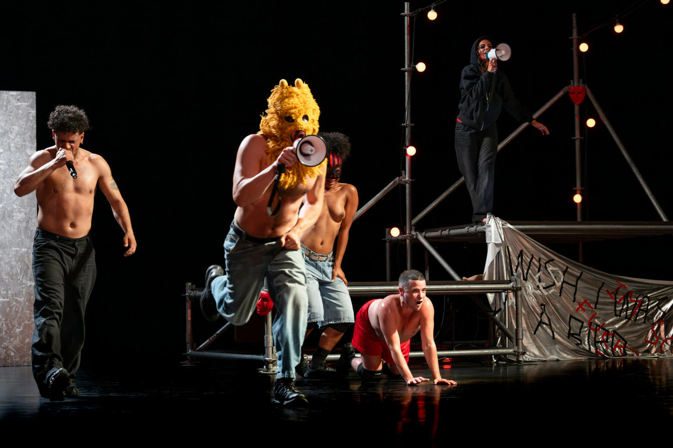 Eine Szene aus Delirious Night: Vier Performer*innen, oberkörperfrei, auf der Bühne, auf der ein Gerüst steht. Zwei von ihnen haben ein Mikro in der Hand. Im Bildvordergrund ein*e maskierte*r Performer*in mit Lautsprecher in der Hand.