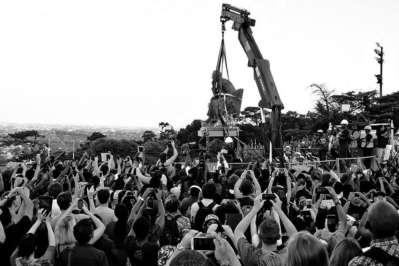 Eine Menschenmenge sieht der Entfernung der Cecile Rhodes Statue an der Universität Kapstadt zu, die nach Protesten abgebaut wurde. Im Mittelpunkt des Bilder hängt die Statue an den Seilen eines Krans, viele Menschen sehen zu.