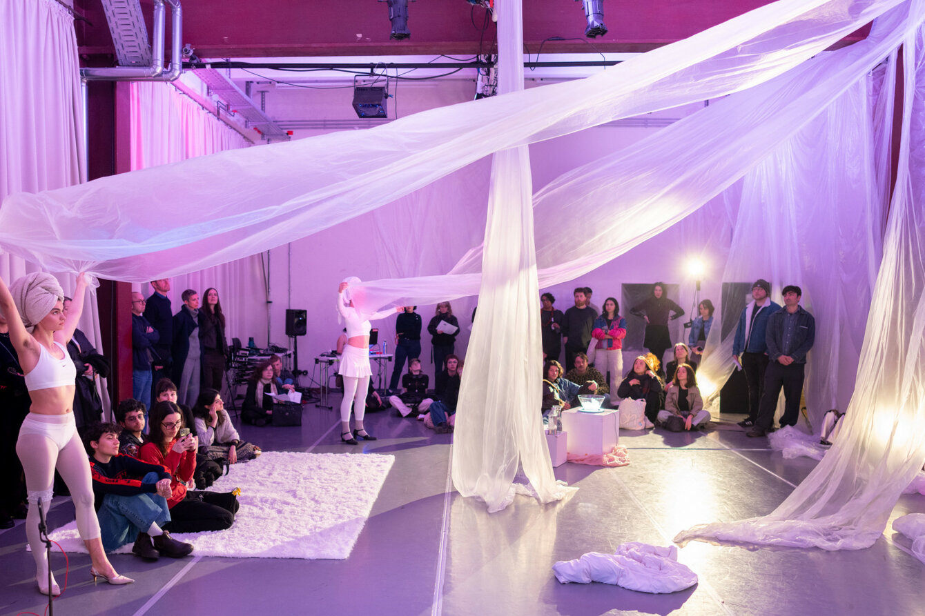 Eine Performance-Situation bei PACT Zollverein: Zwei Performer*innen, jeweils weiß gekleidet stehen auf der linken Seite des Raums, der pink-lila beleuchtet wird und halten weiße Stoffbahnen in der Hand, die oben an der gegenüberliegenden Wand angebracht sind. Das Publikum steht vor den Wänden.