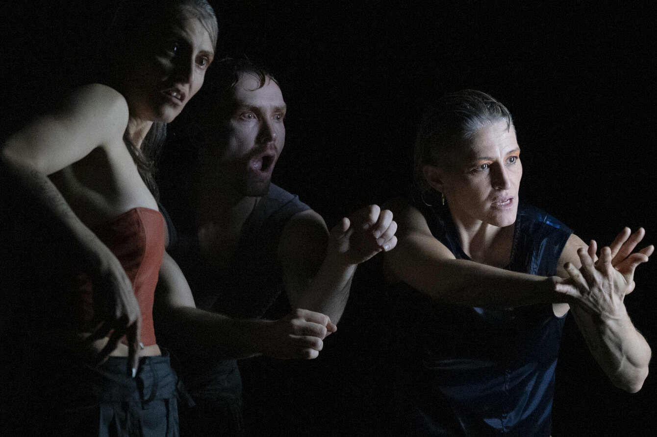 A scene from the performance Mercury Rising: Three people standing in a dark setting in dynamic poses. The person on the left is wearing a sleeveless red top and long black pants. The person in the middle is partially visible and wears a dark top. The person on the right sits on a stool, wearing a sleeveless, dark blue top and olive green pants, arms stretched forward and hands clasped together.