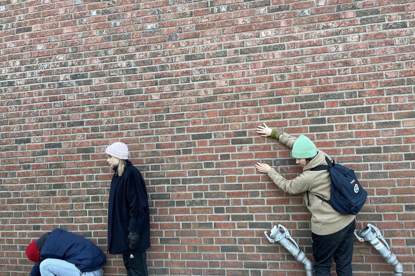 Drei Personen vor einer Ziegelsteinwand bei PACT Zollverein, eine in der Hocke, eine hält sich an den Steinen fest, eine steht. 