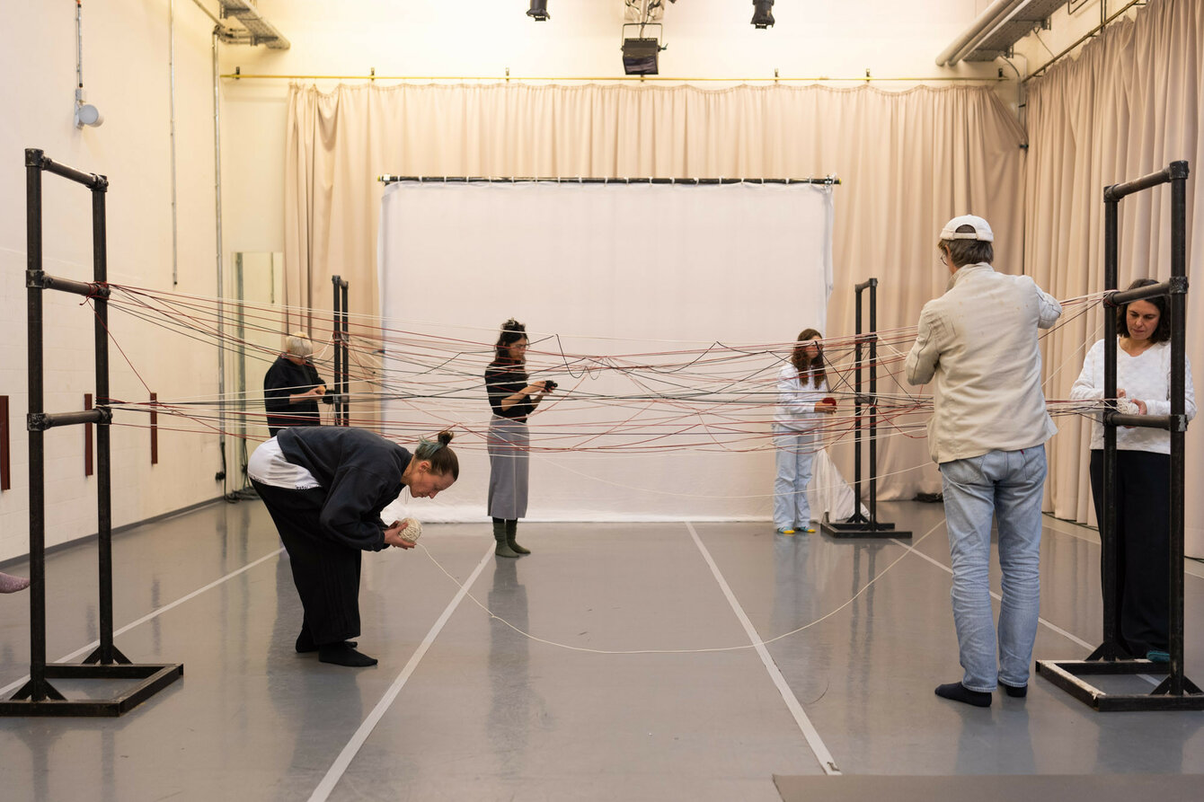 Im Tanzstudio steht eine Installation aus Metallrohren und Fäden die dazwischen gespannt sind. Mehrere Menschen stehen daneben un interagieren mit dem Aufbau. 