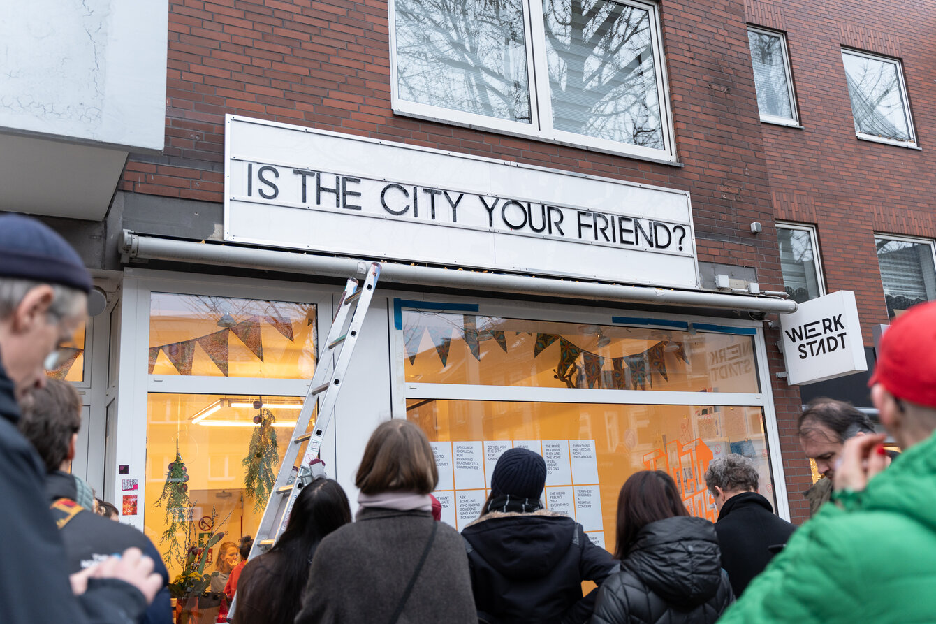 Eine Personengruppe steht vor der WerkStadt in Katernberg und blickt auf die Anzeigetafel über dem erbeutetem Schaufenster. Dort ist in groß angeschlagenen Buchstaben zu lesen "IS THE CITY YOUR FRIEND?"