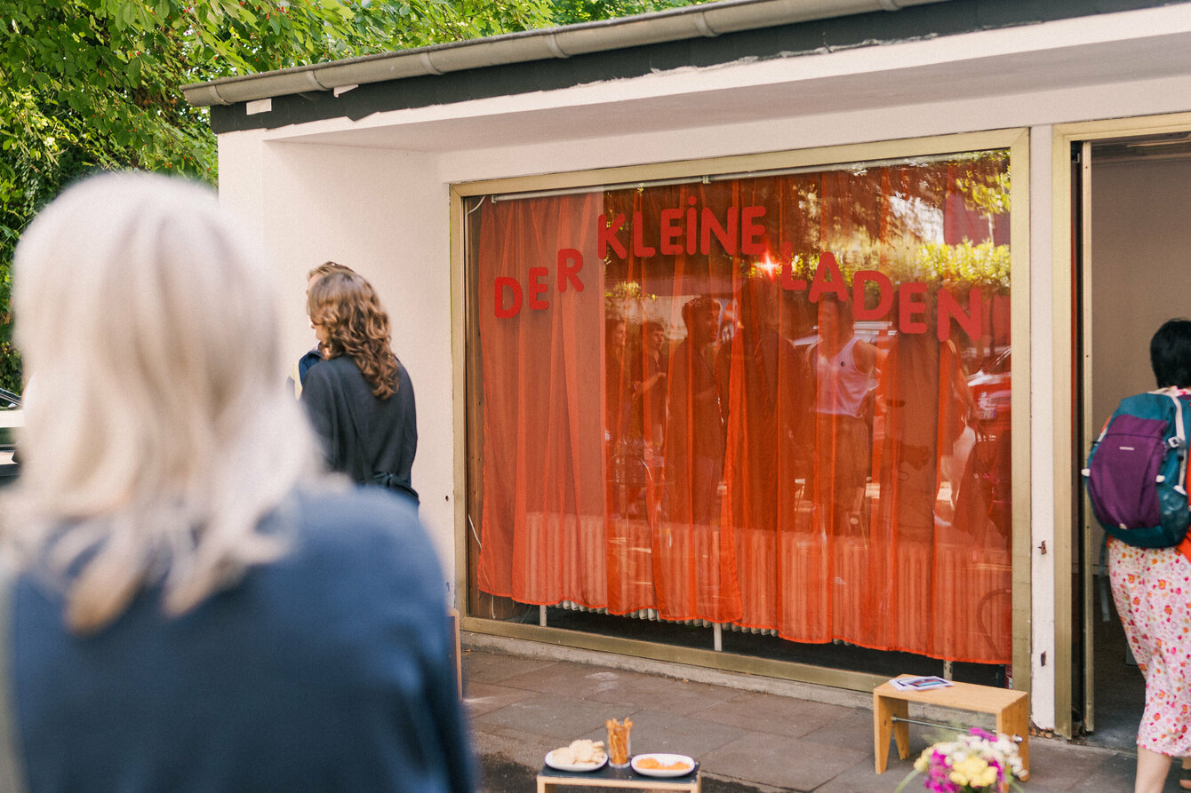 Der Kleine Laden von außen. Es handelt sich um ein kleines Gebäude mit großer Fensterfront auf dem in roter Schrift Der Kleine Laden steht. Hinter der Fensterfront ein roter transparenter Vorhang. Es stehen einige Personen vor dem Kleinen Laden.