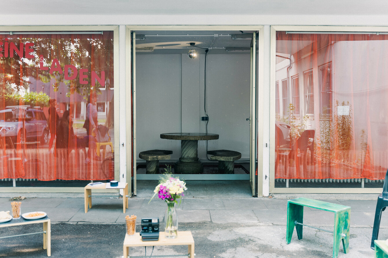Der Kleine Laden von außen. Es zeigt ein kleines Gebäude mit einer großen Fensterfront. Dahinter rote Gardinen. In der Mitte der Fensterfront eine Tür. Sie ist offen, dahinter eine Brunneninstallation. Vor dem Kleinen laden sind kleine Tische zu sehen.