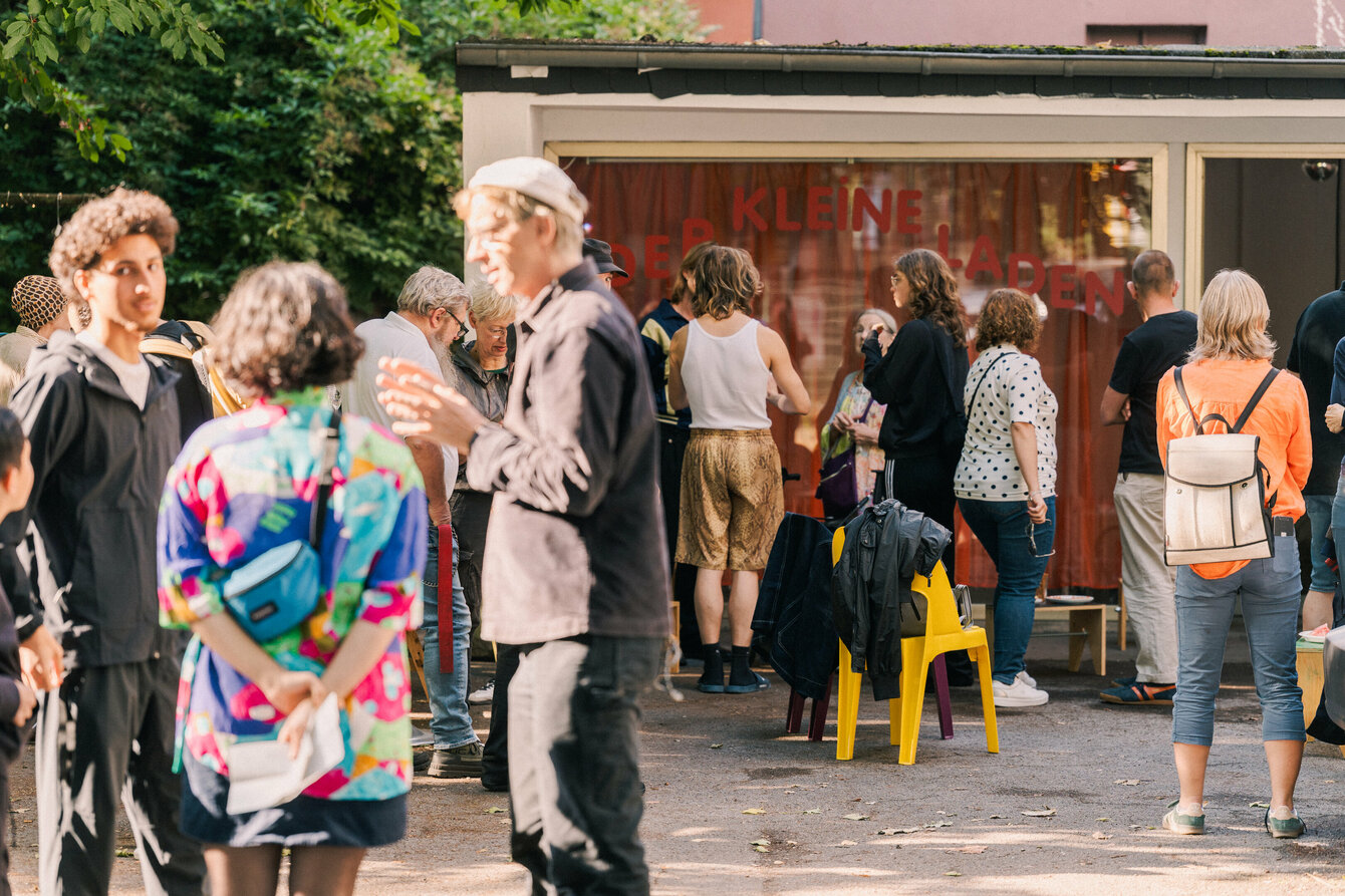 Mehrere Personen sind vor der Fensterfront des Kleinen Ladens zu sehen. Sie stehen in verschiedenen Konstellationen zusammen und unterhalten sich. Es herrscht eine ausgelassene, sommerliche Stimmung, die Abendsonne wirft ein weiches schönes Licht. 
