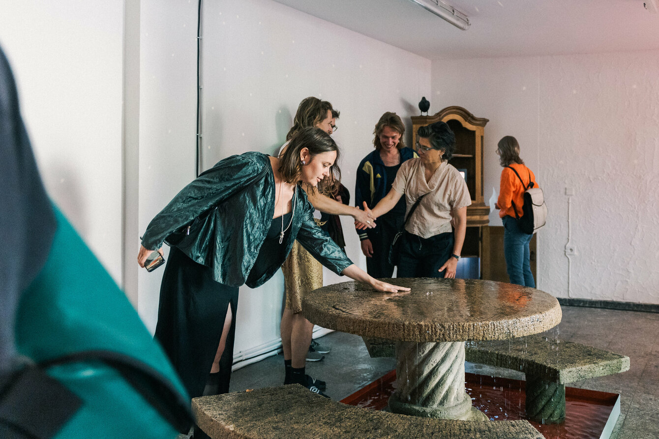 Eine Person im Kleinen Laden steht vor der Installation von Gregor Leiprecht und Konstantin Pütz und streckt die Hand in Richtung Mitte des steinernen Tisches. Dort sprudelt aus einer Öffnung Wasser heraus und läuft über und verteilt sich über den Tisch und das Sitzmobiliar. Ein darunter liegendes Becken fängt das Wasser auf. Im Hintergrund sind weitere Menschen zu sehen, welche sich unterhalten.