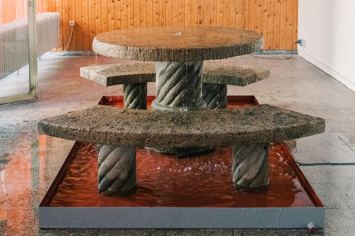Detaillierte Ausstellungsansicht der Installation im Kleinen Laden: Diese besteht aus einem Wasserbecken, in welchem zwei steinerne Bänke und ein steinerner Tisch aufgestellt sind. Aus der Mitte des Tisches sprudelt aus einer Öffnung Wasser über und verteilt sich über die Fläche und das Sitzmobiliar, bevor es im Becken aufgefangen wird. 