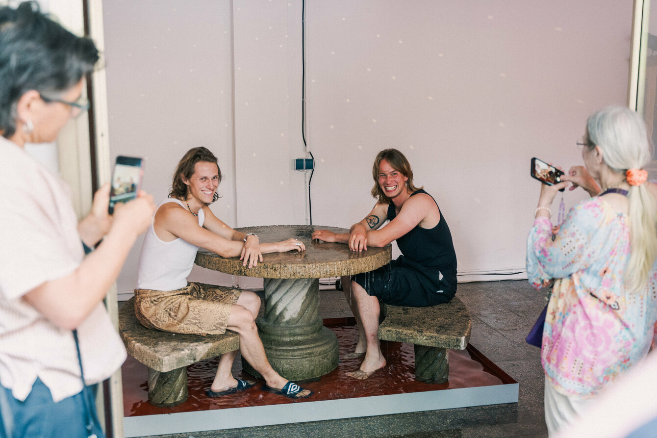 Gregor Leiprecht und Konstantin Pütz sitzen gut gelaunt und lachend im Kleinen Laden an ihrer interaktiven Installation. Diese besteht aus einem Wasserbecken, in welchem zwei steinerne Bänke und ein steinerner Tisch aufgestellt sind. Aus der Mitte des Tisches sprudelt aus einer Öffnung Wasser über und verteilt sich über die Fläche und das Sitzmobiliar, bevor es im Becken aufgefangen wird. 