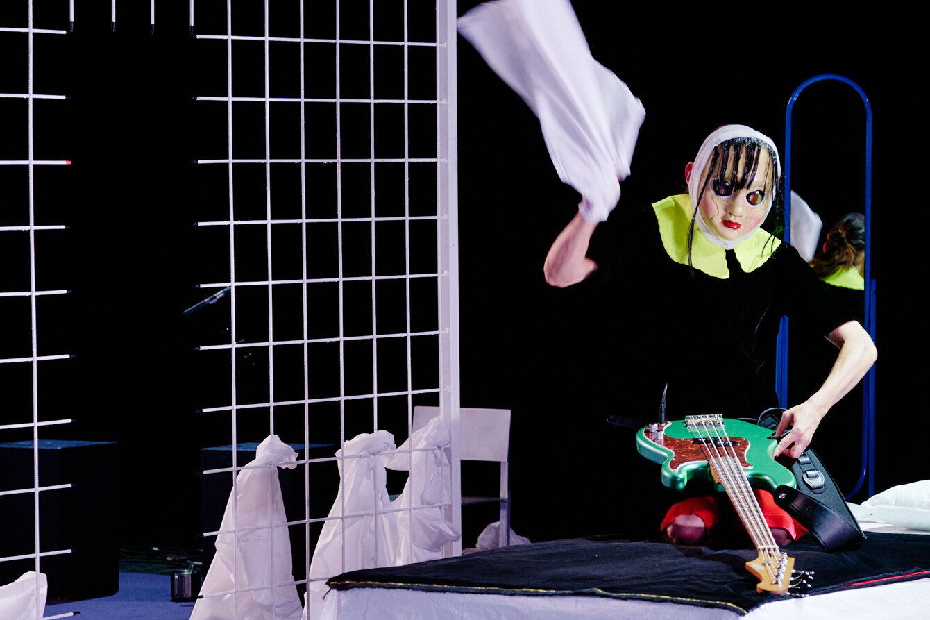 Eine Szene aus Nôt. Ein*e maskierte Performer*in auf der Bühne, die mit einem weißen Tuch nach etwas zu schlagen scheint. Vor der Person eine grüne E-Gitarre. Es handelt sich um eine Art Puppenmaske. Links neben dem*der Performer*in ein weißes Gitter.
