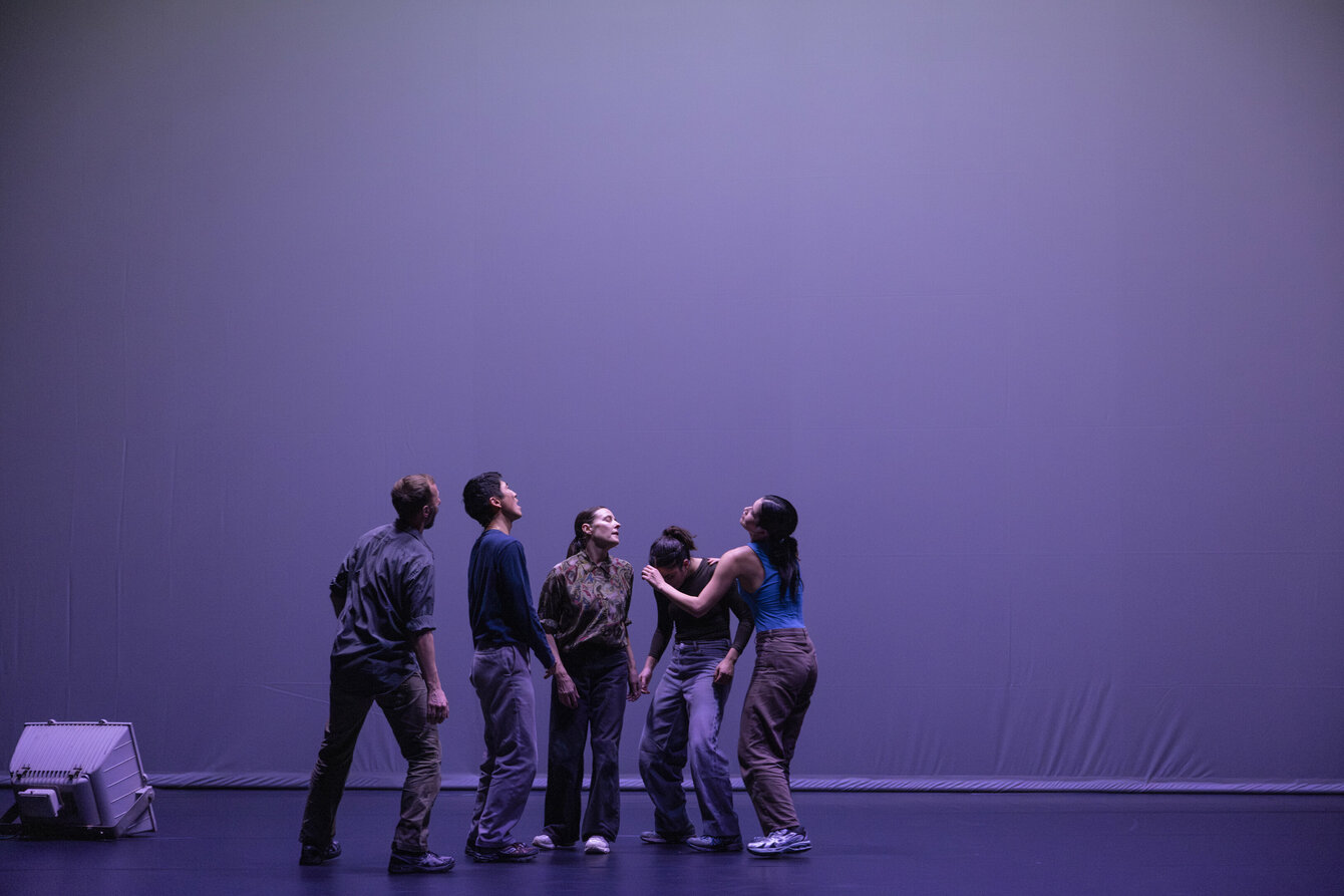 Das Foto zeigt eine Gruppe von Performer:innen, welche zueinander gewandt auf einer Bühne stehen. Sie stehen aufrecht, sind jedoch in Bewegung. Sie sind in weiches Licht gehüllt und verschwimmen mit dem grau-violetten Hintergrund.