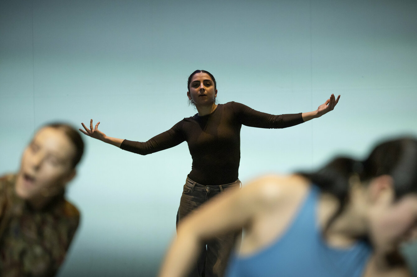 Das Foto zeigt drei Performer:innen auf einer Bühne, wobei zwei von ihnen nur angeschnitten und unscharf im Vordegrund zu erkennen sind. In der Bildmitte und im Fokus der Fotografie schaut eine Tänzer:in frontal in die Kamera streckt die Arme zur Seite aus. Der Hintergrund ist hell, die Performenden setzen sich deutlich von ihm ab. 