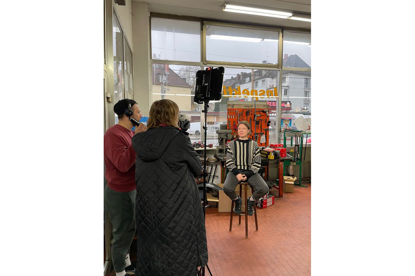 Ein Person sitzt auf einem Hocker in einer Autowerkstatt. Sie wird interviewt und dabei mit einer Kamera gefilmt.
