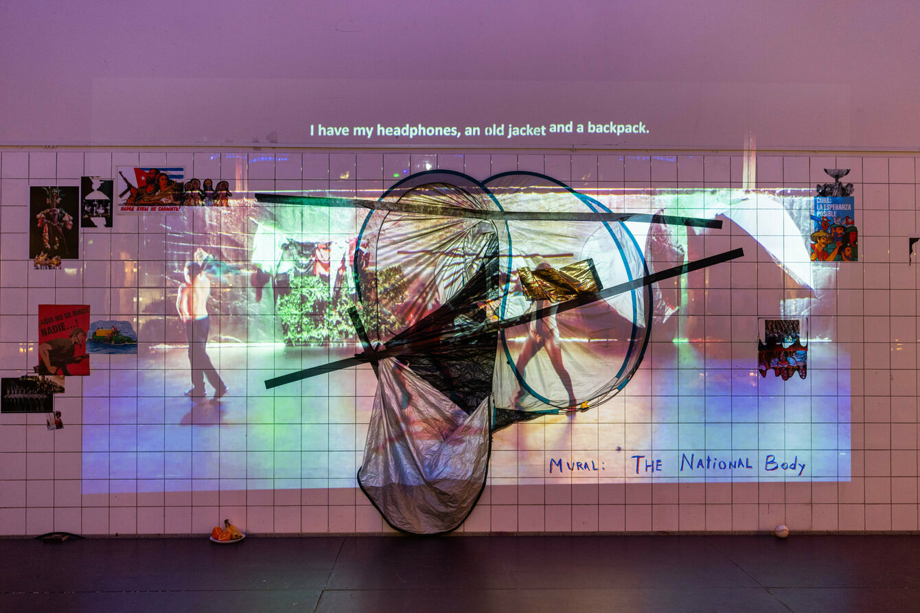 Eine Kunstinstallation die an der Wand befestigt ist. Sie besteht aus einem Zelt, welches mit Gaffertape angeklebt ist und mehreren Fotografien die drumherum aufgeklebt sind. Darüber und überlappend ist ein Video projiziert.