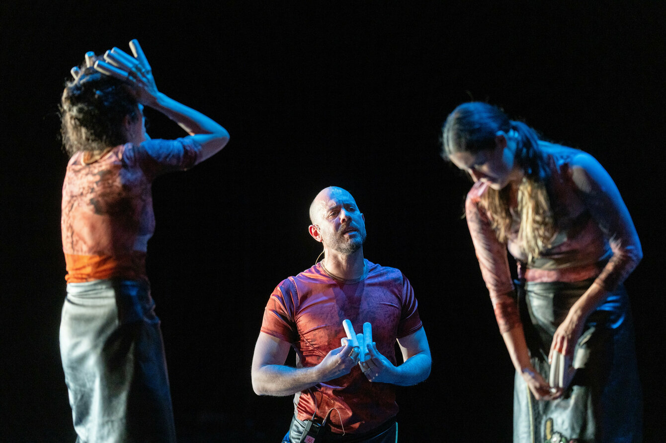 Drei Performer:innen in einer theatralen Blackbox von blauem und weißem Licht beschienen, welches sich mit den rot- und orangefarbenen marmorierten Outfits mischt. Sie stehen separiert von einander in unterschiedlichen Höhen in unterschiedlichen Posen und machen von ihrer Stimme gebrauch.